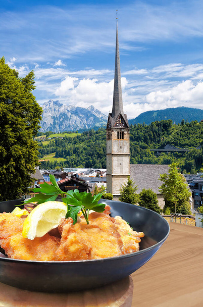 La gastronomía tradicional austríaca (Schnitzel) contra Bischofshofen con la iglesia en Austria - Foto, Imagen
