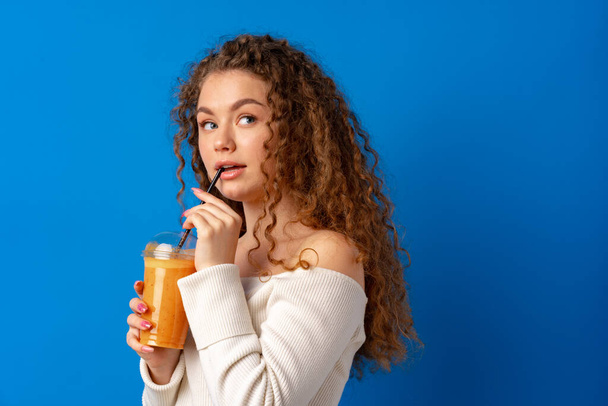 Beautiful curly-haired woman drinking orange juice against blue background - Foto, Imagen
