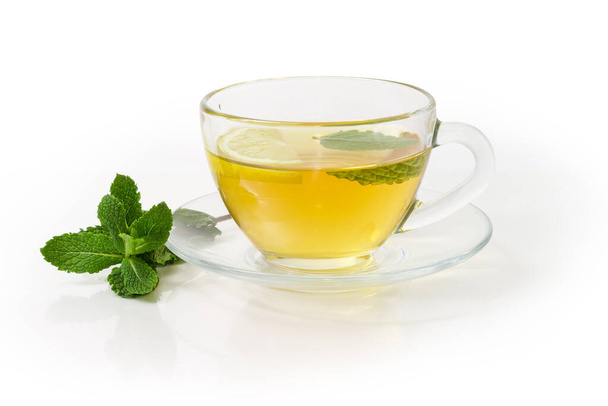 Стеклянная чашка чая с мятным листом и ломтиком лимона на стеклянной тарелке, веточка свежей мяты на белом фоне - Фото, изображение