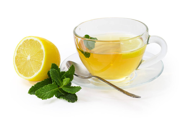 Скляна чашка чаю з листям м'яти та лимонним шматочком на скляній тарілці з чайною ложкою, гілочкою свіжої м'яти та половиною лимона на білому тлі
 - Фото, зображення