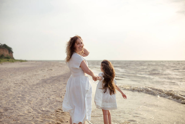 Ganzkörper fröhliche Frau in weißem Kleid umarmt Mädchen, die am Strand in der Nähe des winkenden Meeres am Morgen herumlaufen - Foto, Bild