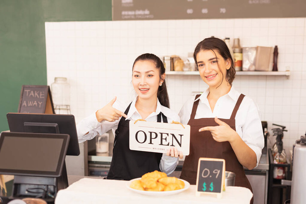 Bal oldalon gyönyörű kaukázusi és ázsiai barista nők mutatják nyitott bolt táblák a kávézó kávézó. Barista munka a kávézóban és a vendéglátóiparban - Fotó, kép