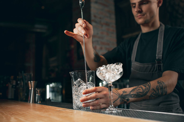 Young caucasian guy, professional bartender in apron mixing cocktails in pub or cafe. - Фото, изображение