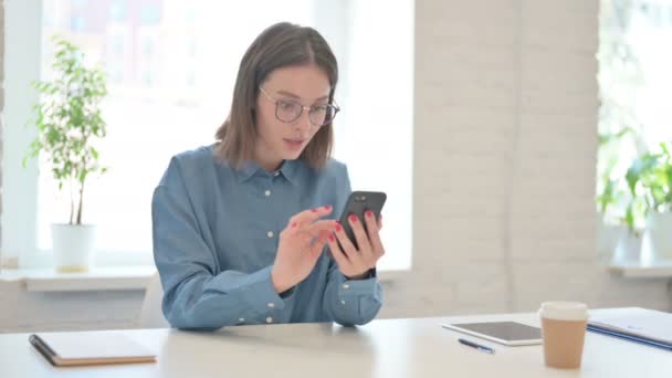 Νεαρή γυναίκα γιορτάζει την επιτυχία στο Smartphone στο γραφείο  - Πλάνα, βίντεο