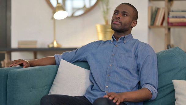 ソファに座っている幸せな若いアフリカ人男性｜Thinking  - 写真・画像