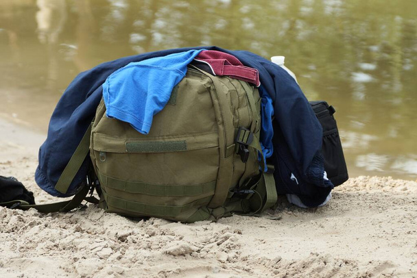un sac à dos vert de l'armée et un tas de vêtements colorés se tient sur le sable sur la plage au bord de l'eau - Photo, image