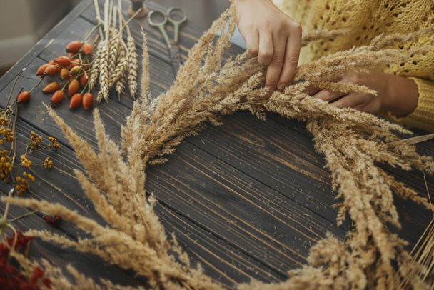 Making rustic autumn boho wreath. Hands holding dry grass and making stylish wreath with wildflowers, herbs and berries on rustic wooden background. Holiday workshop - Фото, изображение