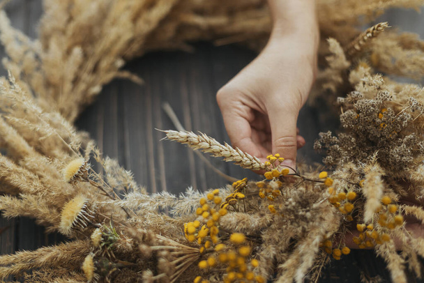 Hands making stylish autumn rustic wreath with dry grass, wildflowers and wheat on rustic wooden table in room. Fall holiday workshop. Florist making boho wreath on dark wood - 写真・画像