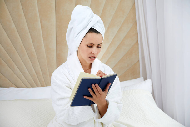 Young woman sits on bed wearing bathrobe writing notes in diary. Blogger creats publication for blog in notebook - Foto, Imagem