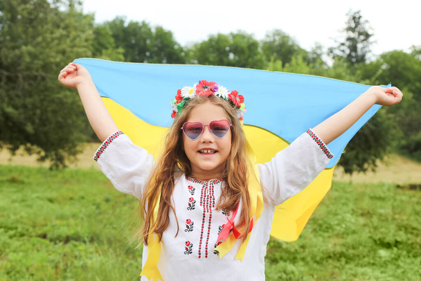 Die gelb-blaue Fahne der Ukraine in den Händen eines schönen Mädchens in einem bestickten Hemd und einem Kranz mit Schleifen. Kind in einem grünen Garten. Unabhängigkeitstag der Ukraine, Tag der Flagge, Verfassung, Bestickte Hemden - Foto, Bild