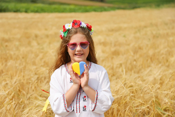 Yellow and blue heart in the hands of a beautiful girl in an embroidered shirt and a wreath with ribbons. Child in a wheat field. Independence Day of Ukraine, Flag Day, Constitution, Embroidery - Photo, image