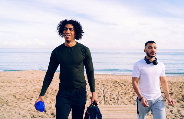 Happy young multiethnic sportsmen in sportswear with bottle of water and headphones communicating and walking away together after workout on seashore - 写真・画像