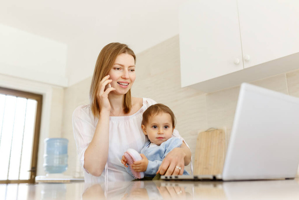 Beautiful business mom is talking on the mobile phone and taking notes while spending time with her cute baby boy at kitchen home office. Working mother concept,Laptop on table. - 写真・画像