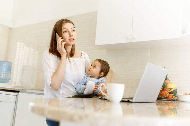 Beautiful business mom is talking on the mobile phone and taking notes while spending time with her cute baby boy at kitchen home office. Working mother concept,Laptop on table. - Fotografie, Obrázek