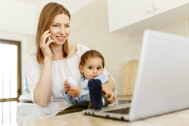 Beautiful business mom is talking on the mobile phone and taking notes while spending time with her cute baby boy at kitchen home office. Working mother concept,Laptop on table. - Foto, afbeelding