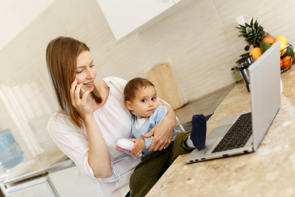 Die schöne Business-Mama telefoniert mit dem Handy und macht sich Notizen, während sie Zeit mit ihrem süßen Baby im Home Office in der Küche verbringt. Working Mother Konzept, Laptop auf dem Tisch. - Foto, Bild