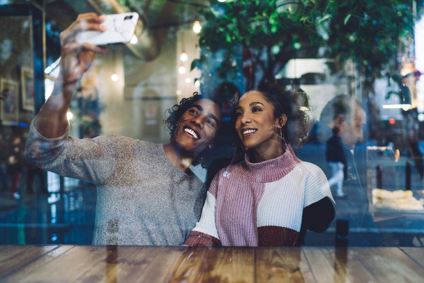 Through glass of cheerful ethnic couple in casual clothes sitting at wooden table in modern cafe and taking selfie on smartphone - Fotoğraf, Görsel