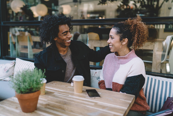 Cheerful multiethnic boyfriend and girlfriend smiling candidly to each other while sitting at small table with coffee to go in modern cafe - Fotografie, Obrázek