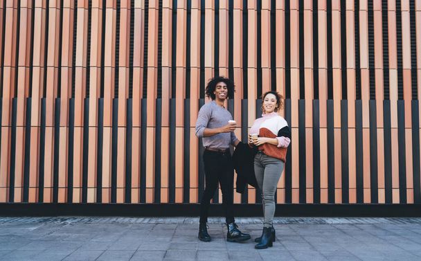 Full length of positive African American couple in casual clothes standing near modern building and drinking coffee while talking and looking at camera - Foto, afbeelding