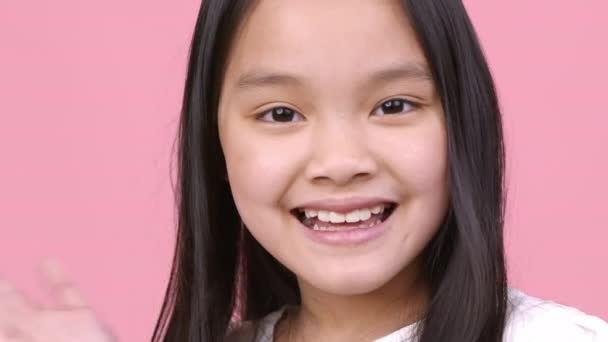 Cheerful greeting. Positive little asian girl waving hand hello and smiling to camera, pink studio background, close up - Footage, Video