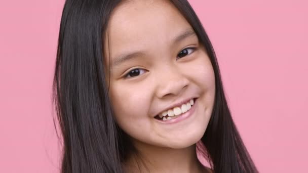 Ευτυχισμένη παιδική ηλικία. Αξιολάτρευτο χαρούμενο κοριτσάκι από την Ασία χαμογελά ευρέως στην κάμερα, ποζάροντας πάνω από ροζ φόντο στούντιο - Πλάνα, βίντεο