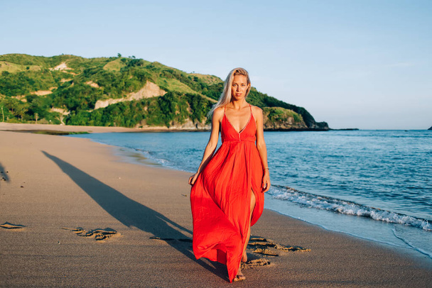 Attractive relaxed carefree woman in bright red sundress strolling along peaceful sandy beach by blue sea and green hilly shore - Valokuva, kuva