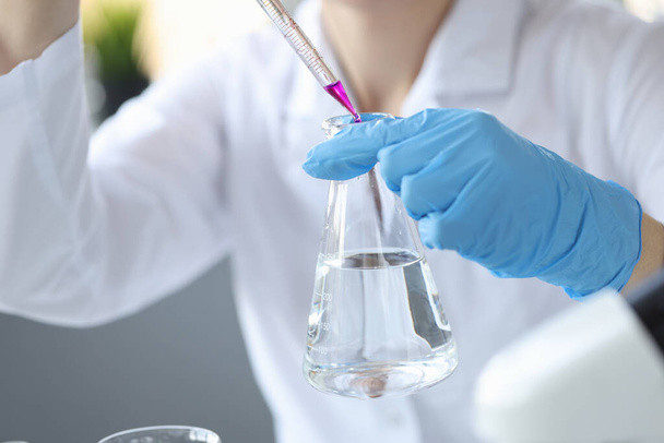Ученый в перчатках держит фляжку с водой и капает в нее фиолетовую жидкость из пипетки. - Фото, изображение