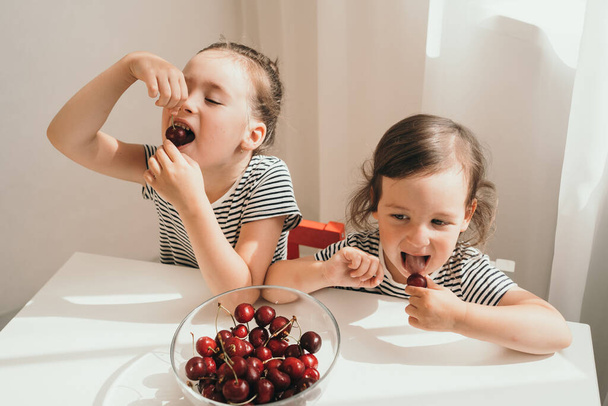 幸せな子供たちは夏の果実を食べる。おいしい明るいジューシーなベリー。縞模様のTシャツの子供たちが食べる。二人の姉妹は午後の軽食のためにチェリーを食べる。子供たちはテーブルに座って食べます - 写真・画像