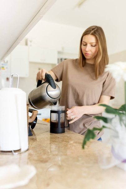 Beautiful young woman sanding at kitchen counter, pouring herself a cup of hot coffee, enjoying leisure time at home in the morning. - Photo, Image