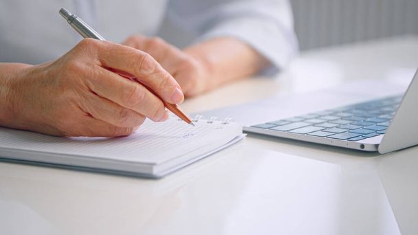 Стара жінка зморшкуваті руки пишуть у паперовому блокноті з ручкою за столом біля сірої клавіатури ноутбука
 - Фото, зображення