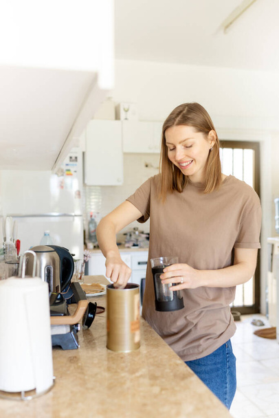 Happy young woman making coffee in kitchen - french press coffee. Caucasian female model in her twenties at home kitchen. Lifestyle concept. - Photo, image