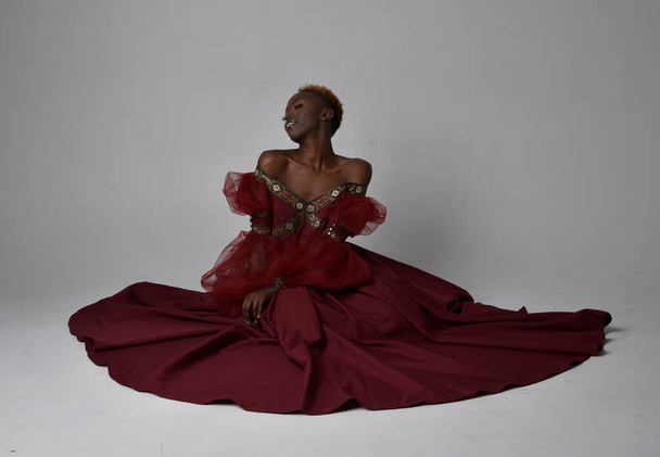 Full length portrait of pretty African woman wearing long red renaissance medieval fantasy gown, sitting pose on a light grey studio background. - Photo, Image