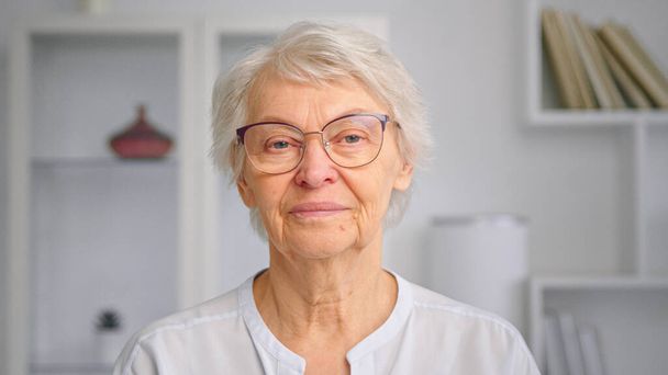短い灰色の髪と眼鏡を持つ高齢者の女性はまっすぐ見えると笑顔陽気に白い家具に対して立って - 写真・画像