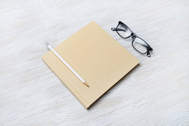 Foto eines geschlossenen, quadratischen Buches mit Brille und Bleistift auf hellem Holzhintergrund. Vorlage für die Platzierung Ihres Designs. - Foto, Bild