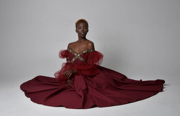 Πλήρες πορτρέτο της όμορφης Αφρικανής γυναίκας που φοράει μακρύ κόκκινο φόρεμα της αναγέννησης μεσαιωνικό φαντασία, κάθεται θέτουν σε ένα ανοιχτό γκρι φόντο στούντιο. - Φωτογραφία, εικόνα