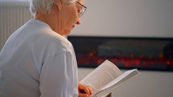 Pensioner lady teacher in glasses sits on large grey sofa and reads book against artificial fireplace at home - Photo, image