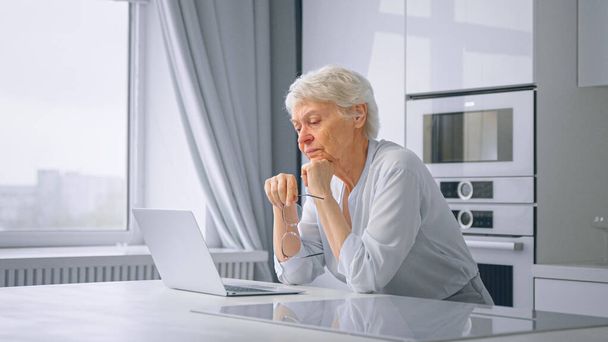 Продумана старша леді секретарка з коротким волоссям тримає окуляри в руці і сидить в сірому ноутбуці на кухні
 - Фото, зображення