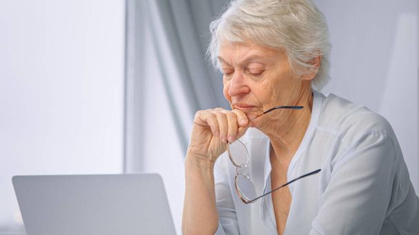 Ragionevole segretaria anziana con i capelli corti tiene gli occhiali in mano e si siede al computer portatile grigio in cucina a casa - Foto, immagini