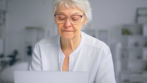 Donna d'affari anziana concentrata in tipi di occhiali su computer portatile e sorride allegramente seduto a casa contro mobili - Foto, immagini