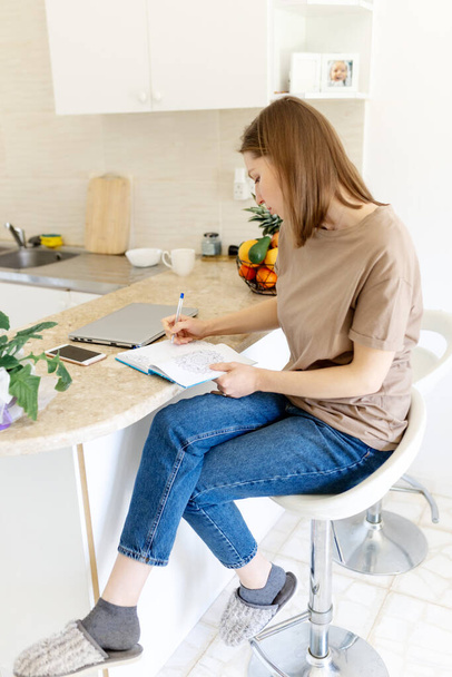 Jolie jeune femme prenant des notes à la maison sur un bureau en bois dans la cuisine. Intérieur de la maison, fille étudiant, écriture sur papier. - Photo, image