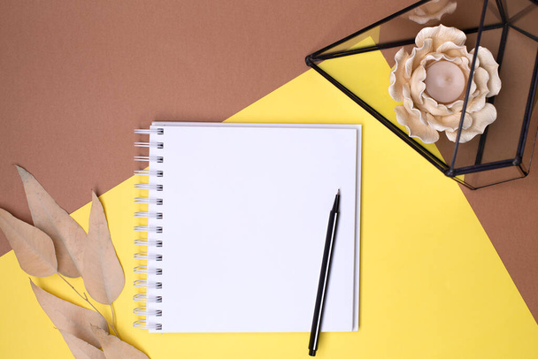 Top view of an empty diary with a spring and a pen, beige leaves, glass florarium with a candle in the shape of a flower on a combined yellow-brown background, place for text - Fotó, kép