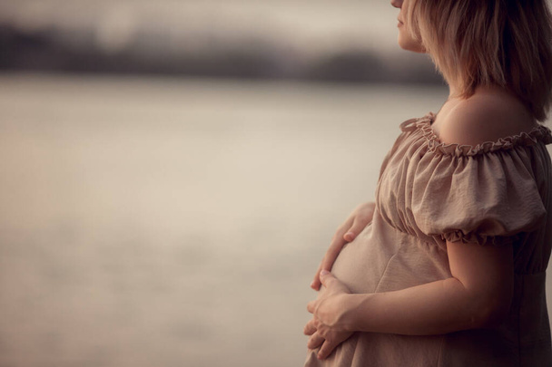 těhotná žena v béžových šatech drží ruku na bříšku. Čekám na dítě, těhotné focení. těhotenství a porod v létě, 9 měsíců. mateřství koncept - Fotografie, Obrázek