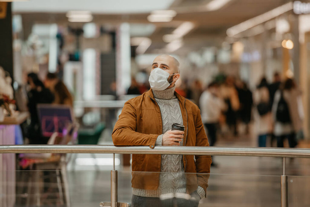 Un hombre con una máscara facial para evitar la propagación del coronavirus está sosteniendo una taza de café mientras espera en el centro comercial. Un tipo calvo con una máscara quirúrgica mantiene una distancia social.. - Foto, imagen
