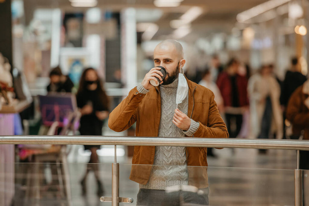 Bir adam alışveriş merkezinde kahve içerken tıbbi maske takıyor. Kel bir adam sosyal mesafeyi koruyor.. - Fotoğraf, Görsel