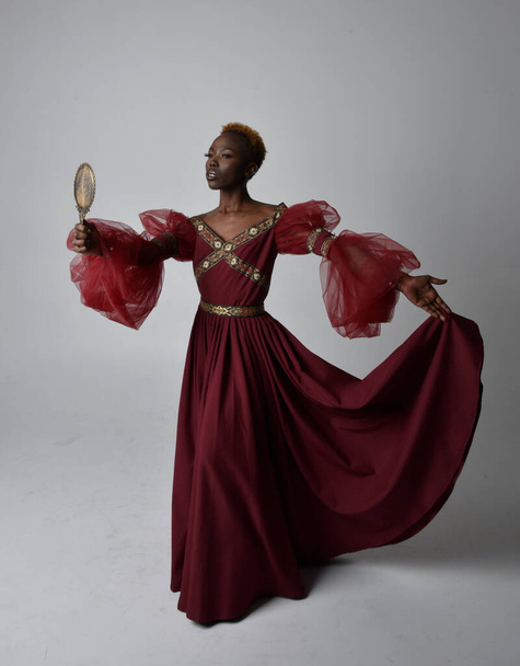 Full length portret van mooie Afrikaanse vrouw dragen lange rode renaissance middeleeuwse fantasie toga, staande pose op een lichtgrijze studio achtergrond. - Foto, afbeelding