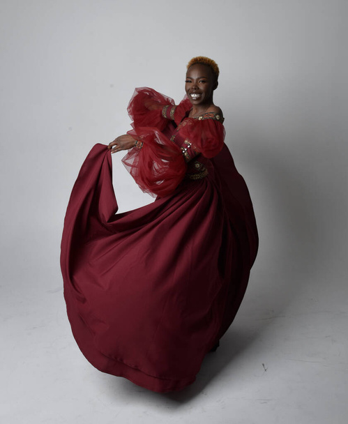 Portrait pleine longueur de jolie femme africaine portant une longue robe de fantaisie médiévale renaissance rouge, pose debout sur un fond de studio gris clair. - Photo, image