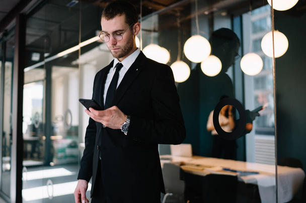 Confident businessman in formal wear and glasses standing in office and messaging on mobile phone while having break during work - Fotografie, Obrázek
