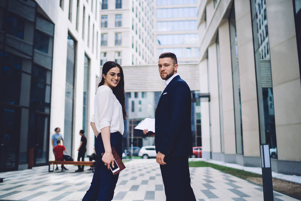 Side view of smiling young confident business employees in formal clothes standing against modern skyscraper and talking at camera during daytime break - Foto, immagini
