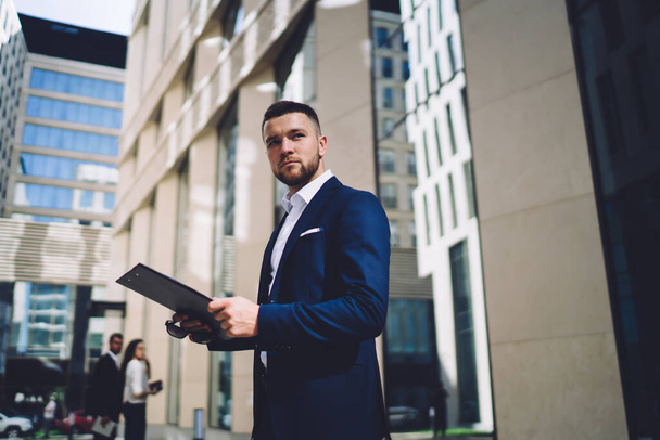 Low angle of serious young businessman in formal wear looking away while standing by modern building with clipboard in hands - 写真・画像