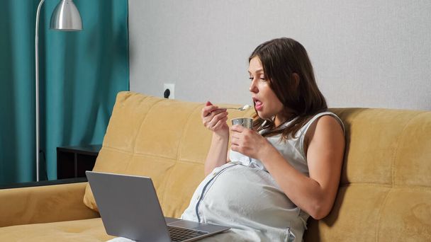 Young pregnant woman eating yogurt and looking at laptop while sitting on sofa - Fotó, kép
